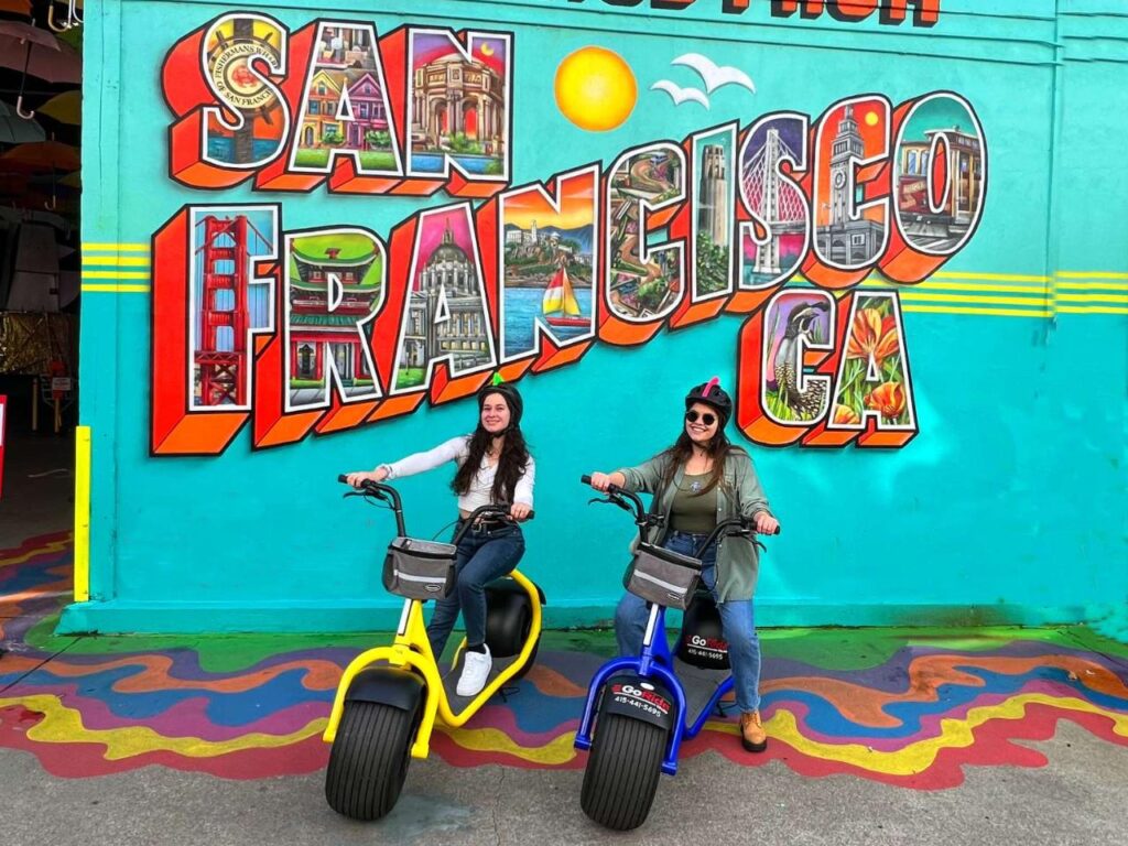 Two women on Scooters in front of San Francisco Mural