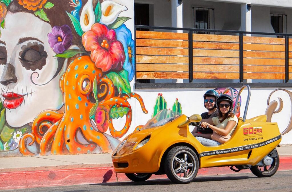 couple in little yellow gocar driving past san diego mural